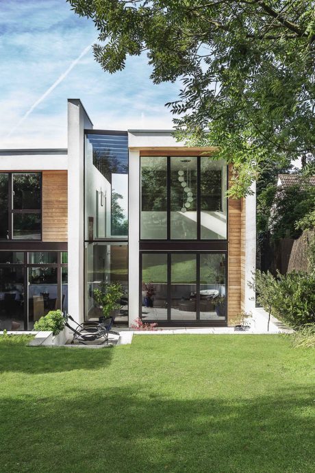 A modern, glass-walled house surrounded by lush greenery and an expansive lawn.