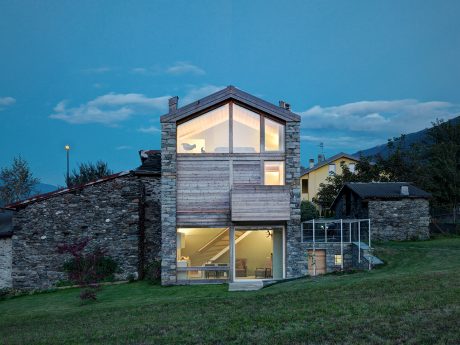 A modern stone and wood home with large windows overlooking a grassy lawn and mountains.