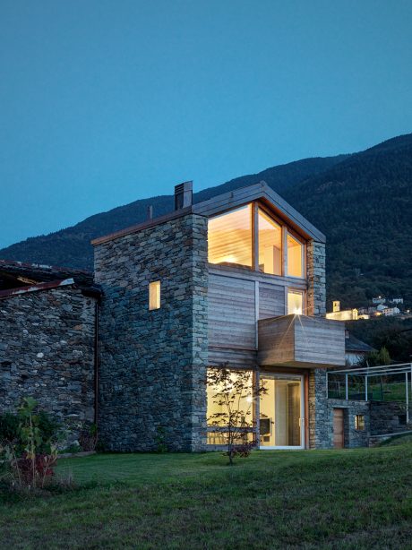 A modern, two-story stone house with wooden accents and large windows overlooking a mountainous landscape.