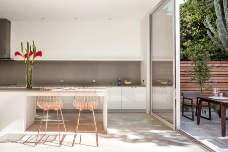 Modern kitchen with sleek grey cabinetry, minimalist copper barstools, and floor-to-ceiling windows overlooking a lush outdoor area.
