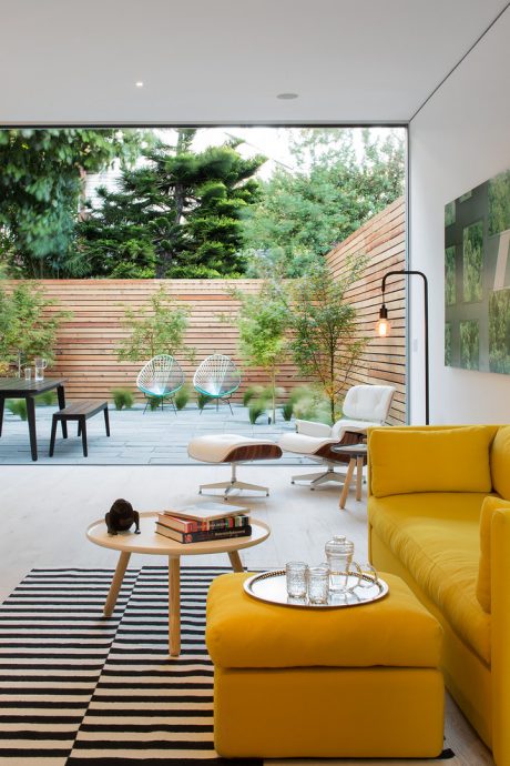 Bright open-plan living space with wood-slat fencing, comfy yellow sofa, and glass doors to garden.