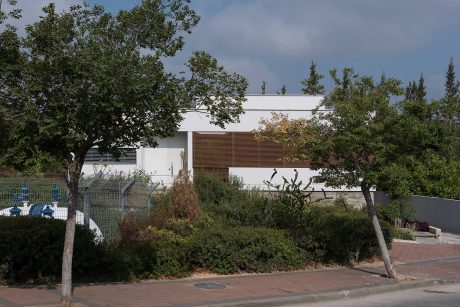 White modern building with wooden slats, surrounded by lush greenery and paved pathway.
