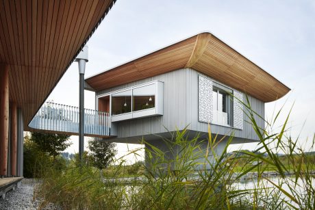 A modern cabin-style home with a prominent wooden roof, glass walls, and lush vegetation.