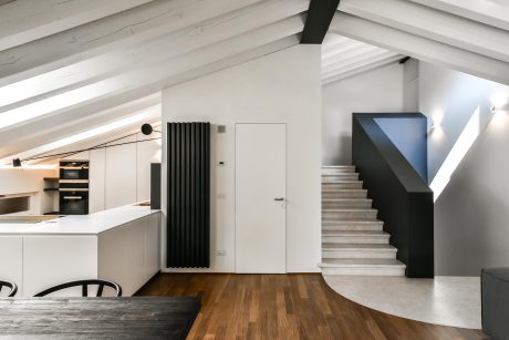 Minimalist kitchen with wooden floors, white walls, and a sleek black radiator. Staircase leads to upper level.