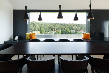 Modern dining room with large black dining table, black chairs, and pendant lights overlooking scenic forest view.