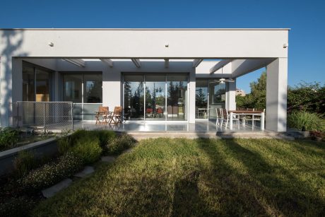 Modern glass-walled home with open patio, lawn, and wooden outdoor furniture.