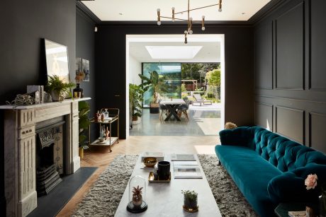 Sleek, dark living area with a tufted blue sofa, ornate fireplace, and lush greenery beyond.