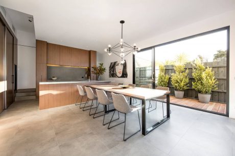 Modern kitchen and dining area with wooden cabinets, large windows, and stylish light fixture.