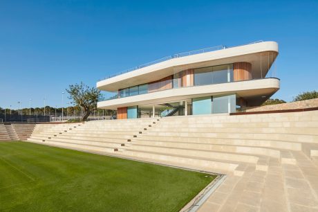 A modern, multi-level building with sleek, curved lines, expansive glass panels, and a grand staircase leading to a grassy field.