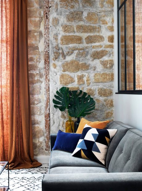 Rustic stone wall, gray sofa with patterned pillows, potted plant, and orange curtains.