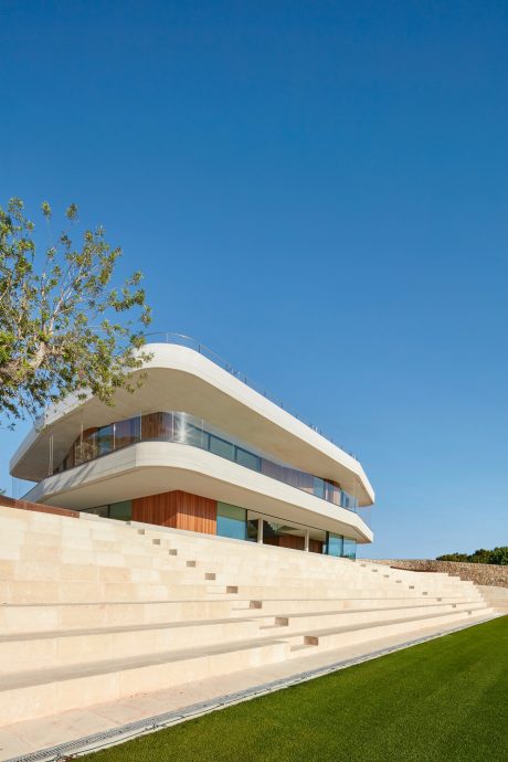 A modern, curved building with glass walls, wooden accents, and a stepped terrace leading to a lawn.