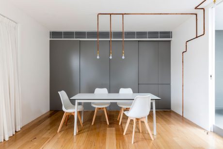 A modern, minimalist dining room with a white table, wooden chairs, and copper pipe lighting fixtures.