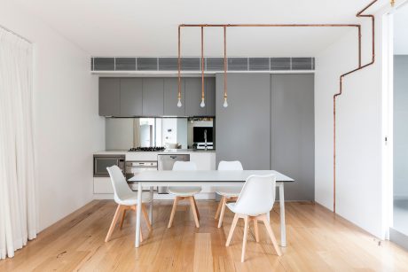 Minimalist kitchen with copper pipe lighting, white dining table and chairs on hardwood floor.