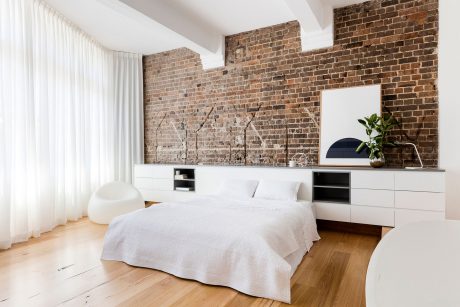 A spacious bedroom with exposed brick walls, white curtains, and a modern dresser.