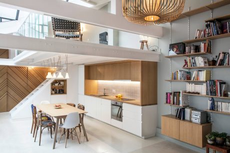 Modern, open-concept kitchen-living room with wooden cabinets, bookshelves, and pendant lighting.