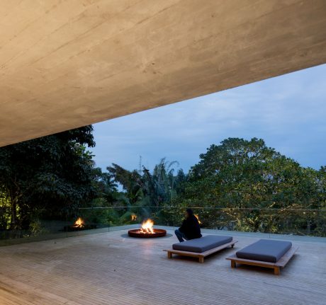 A contemporary outdoor lounge area with a concrete overhang, fire pit, and cushioned benches overlooking lush vegetation.