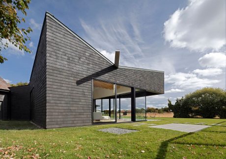 A modern, angular house with large windows, surrounded by a lush lawn and trees.