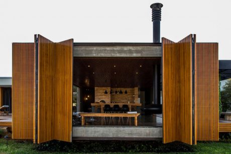 A modern wooden pavilion with a central fireplace and dining area surrounded by nature.