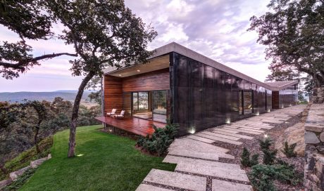 Modern glass-walled structure with wooden elements, surrounded by lush greenery and a paved path.