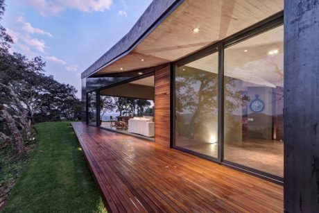 Expansive, glass-enclosed modern home with wood decking and overhanging roof structure.