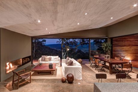 Spacious modern rustic living room with panoramic mountain view, warm lighting, and wooden furnishings.
