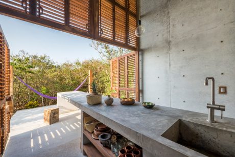 Modern outdoor kitchen with concrete countertops, wooden shutters, and natural vegetation.