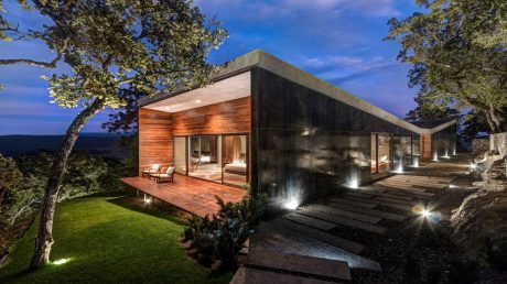 Contemporary wood and concrete home with expansive glass walls and a deck overlooking a wooded landscape.