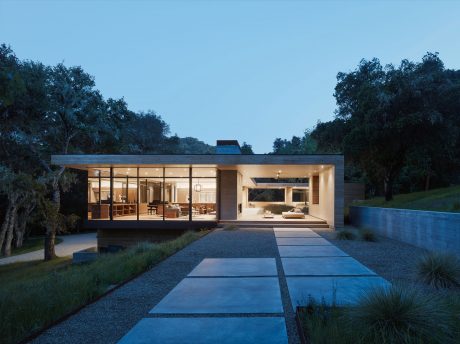 A modern, glass-walled house nestled among trees, with a sleek patio and reflecting pool.