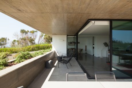 Spacious covered balcony with concrete ceiling and sliding glass doors, surrounded by lush greenery.