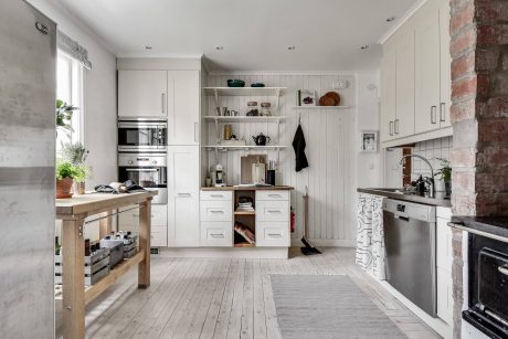 A minimal, bright kitchen with white cabinets, wooden countertops, and an exposed brick wall.
