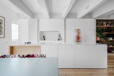 Minimalist kitchen design with white cabinetry, wood accents, and colorful upholstered bench.
