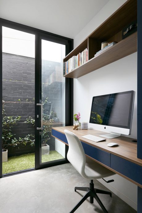A modern home office with sleek wooden shelves, a computer desk, and large glass doors overlooking lush greenery.