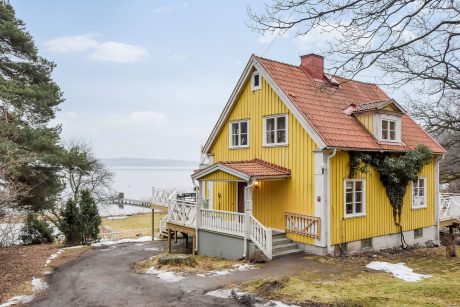 A charming yellow wooden house with a red tiled roof, a porch, and a scenic lakeside view.