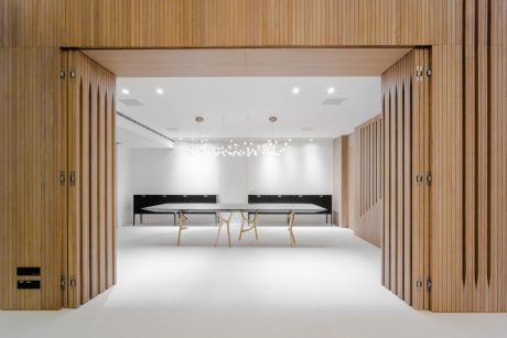 Modern, minimalist interior design featuring a large open space with wooden panels and white walls, recessed lighting, and a long rectangular table with chairs.