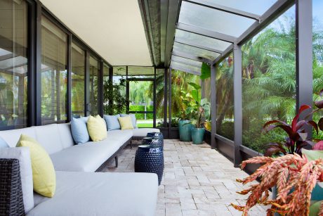 A contemporary sunroom with wicker furniture, potted plants, and a glass roof overlooking a lush garden.