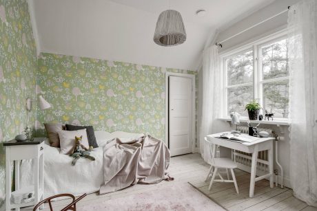 A cozy bedroom with a floral wallpaper, a desk, and a large window overlooking the outdoors.