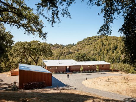A rustic brick building with a metal roof surrounded by lush greenery and a gravel path.