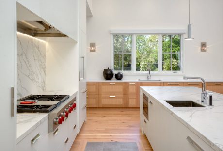 Spacious, modern kitchen featuring wooden cabinetry, marble countertops, and large windows overlooking lush greenery.