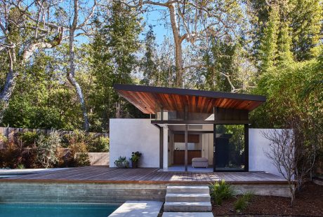 A modern, wooden-roofed pavilion with a glass facade overlooking a pool and wooded landscape.