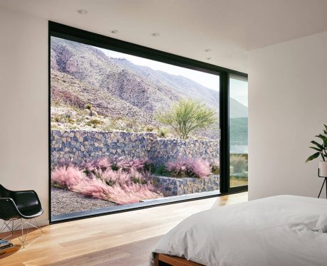 A bedroom with a large picture window overlooking a desert landscape with mountains, a stone wall, and vibrant pink plants.