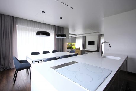Modern, minimalist kitchen and dining area with sleek white countertops, pendant lights, and curtained windows.