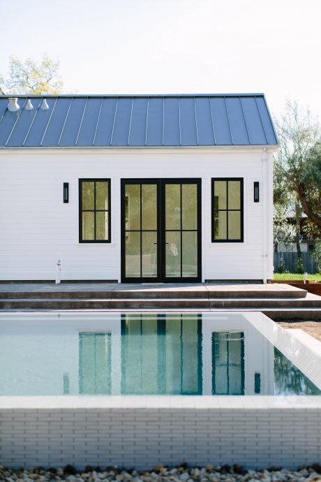 A modern, minimalist building with large glass windows, a metal roof, and a pool surrounded by a brick border.