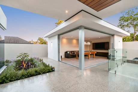 Contemporary open-plan living space with floor-to-ceiling glass walls, wooden ceiling, and lush greenery.