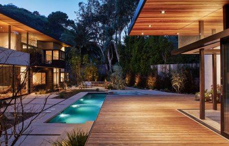 Elegant modern residence with wooden deck, pool, and lush landscaping at dusk.