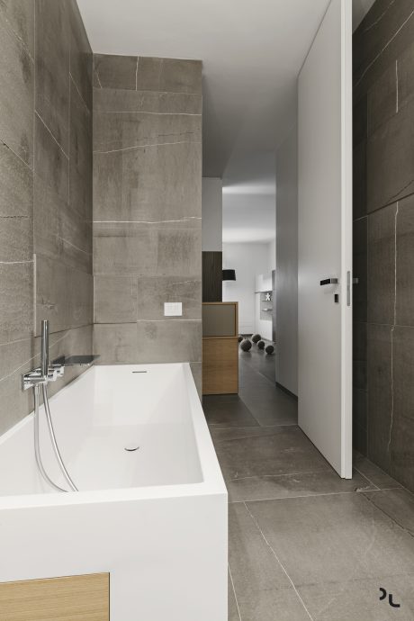 Modern bathroom with minimalist design, featuring a sleek white tub, gray tiled walls, and a glimpse of the adjacent room.