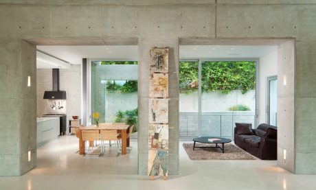 Modern, open-concept interior with concrete walls, large windows overlooking greenery, and a minimalist furniture arrangement.