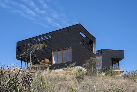 Modern black wooden house with large windows, set against a blue sky and rugged landscape.