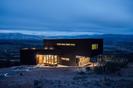 A modern, two-story wooden house with large windows overlooking a mountainous landscape.