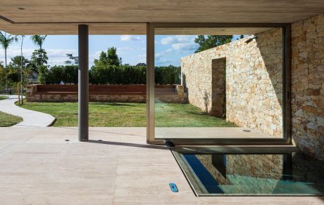 Modern stone architecture with expansive glass windows, reflecting pool, and lush greenery.
