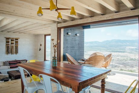 Rustic cabin interior with wooden beams, yellow lamps, and panoramic mountain view.